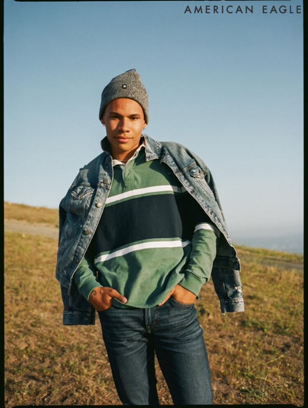 Young man wearing a gray knit beanie, denim jacket and striped polo shirt standing in a field with a blue sky backdrop.