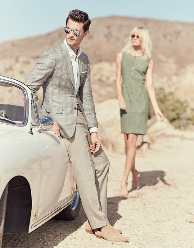 A fashionable couple is posing with a vintage car in an outdoor setting. The man is leaning on the car, and the woman stands nearby.