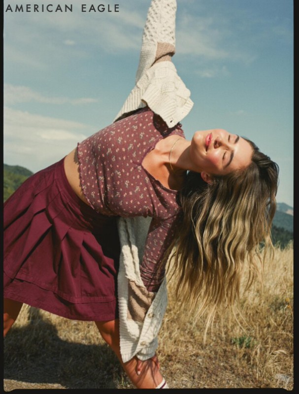 Woman strikes yoga pose outdoors wearing a floral print top, maroon pleated skirt, and a cream-colored cardigan draped over her shoulders.