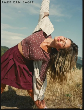 Woman in floral crop top and pleated maroon skirt stretching outdoors under a clear blue sky