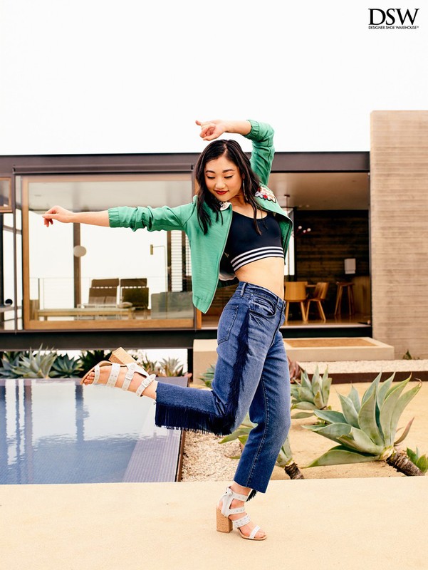 A young woman in jeans, crop top, and green jacket poses dynamically near a modern building and swimming pool, showcasing a fashion-forward look.