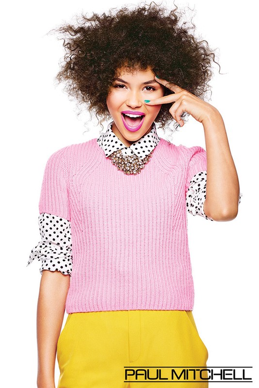 A joyful young woman with an afro poses, wearing a pink knit sweater, yellow pants, and a polka-dot blouse against a white backdrop.
