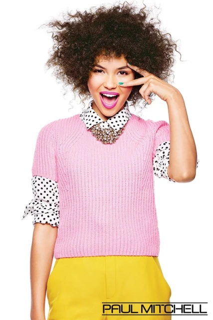 A joyful young woman with an afro poses, wearing a pink knit sweater, yellow pants, and a polka-dot blouse against a white backdrop.