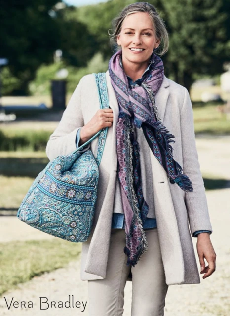 Adult woman wearing a coat and patterned scarf, carrying a patterned tote bag, stands outdoors on a sunny day in natural lighting.