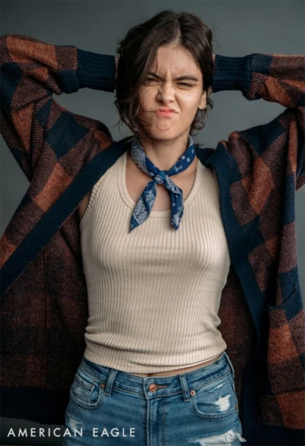 A young woman with dark hair and blue jeans, wearing a cardigan and tank top, puckers her lips in a studio shot against a grey background.