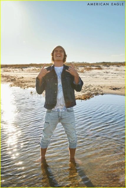 Full shot of an adult man standing in the water at the beach wearing jeans, a t-shirt, and a jacket making a hand gesture.