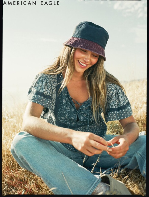 A smiling woman in a bucket hat and floral top sits in a sunny field, wearing jeans, showcasing a casual, outdoor fashion aesthetic.