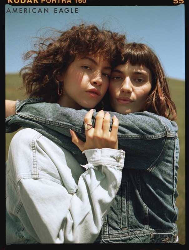 Two women in denim jackets pose outdoors with arms around each other. American Eagle brand is visible. Fashion portrait of young adults.
