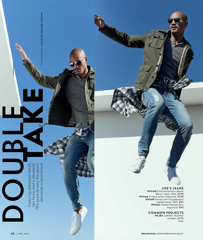 Fashion editorial shot of a male model in olive jacket, jeans, and sneakers, striking a dynamic pose against a blue sky and white geometric background.