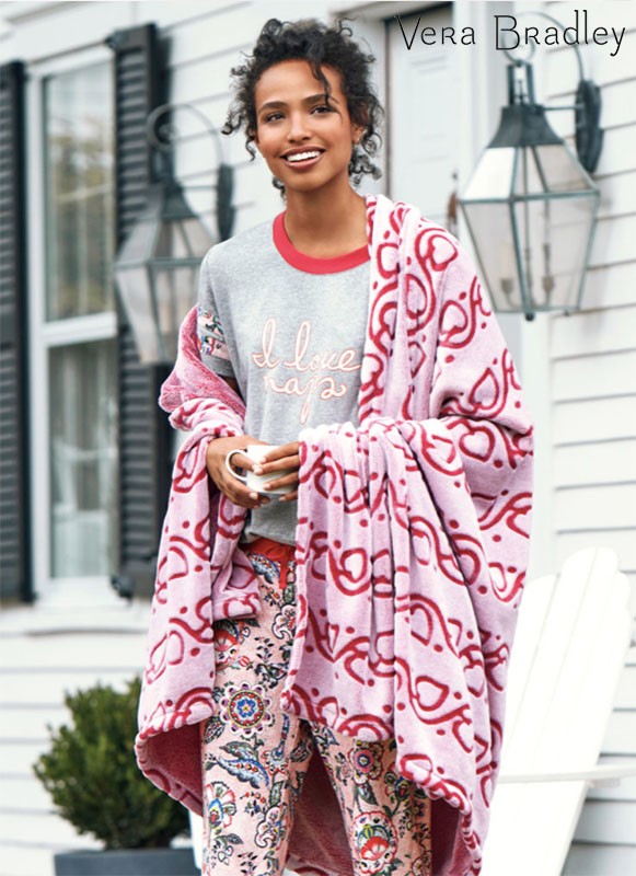 Woman in pajamas and a blanket stands outside a white house with dark shutters, holding a mug in a Vera Bradley product shot.