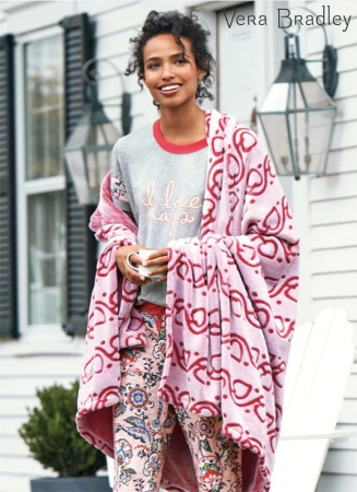 Woman in pajamas and a blanket stands outside a white house with dark shutters, holding a mug in a Vera Bradley product shot.