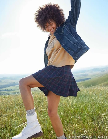 Smiling woman with afro hair wearing a denim jacket, plaid skirt and white boots in a grassy landscape