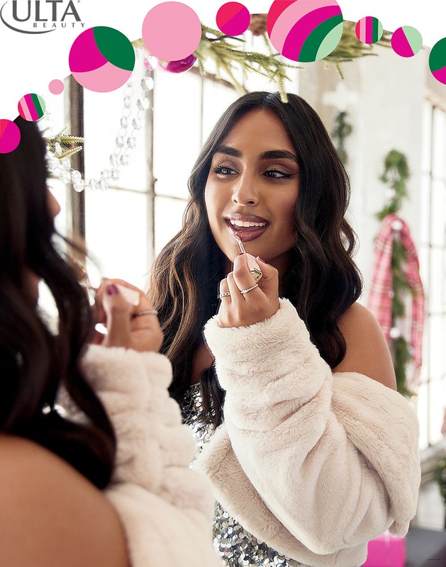 Woman applies lipstick in front of mirror, wearing a white fur coat and a sparkling top, decorated with ornaments and Christmas garlands.