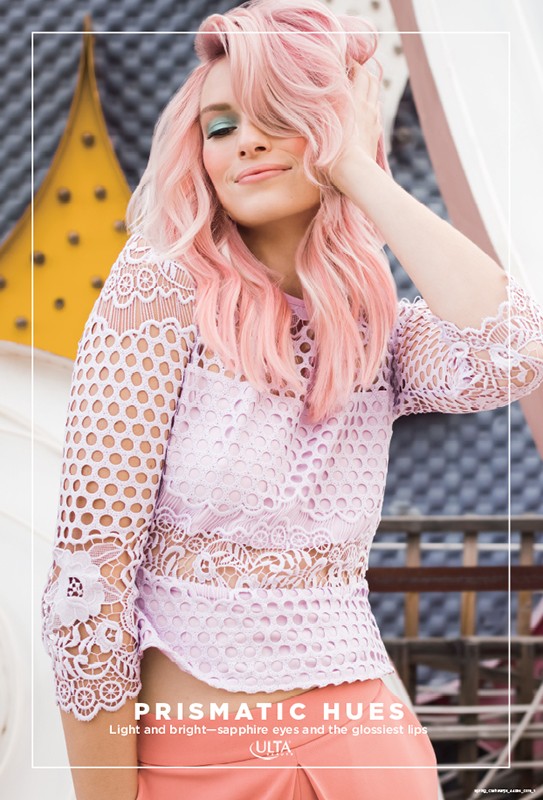Adult female with pink hair and light eye makeup poses in a studio with white background and yellow geometric prop. She is wearing a lace top.