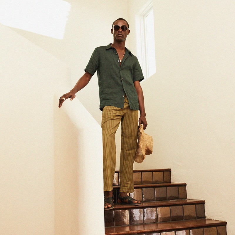 Dark-skinned adult male in casual resort wear posing on stairs. He wears sunglasses, a green shirt, striped pants and holds a straw hat.