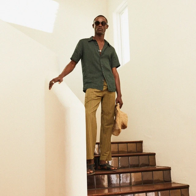 Dark-skinned adult male in casual resort wear posing on stairs. He wears sunglasses, a green shirt, striped pants and holds a straw hat.