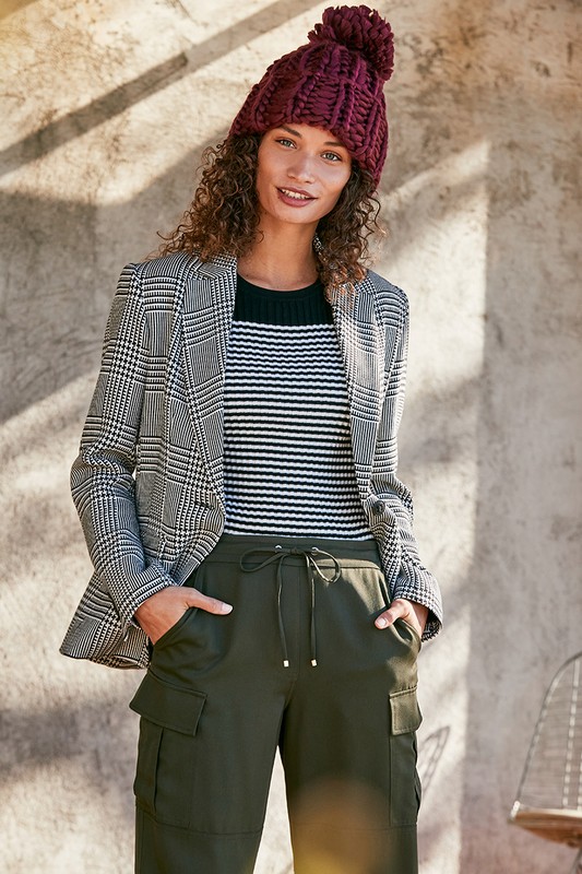 Stylish adult female wearing a plaid blazer, striped top, cargo pants, and burgundy knit hat against a textured neutral wall.