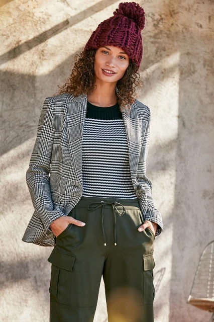 Stylish adult female wearing a plaid blazer, striped top, cargo pants, and burgundy knit hat against a textured neutral wall.