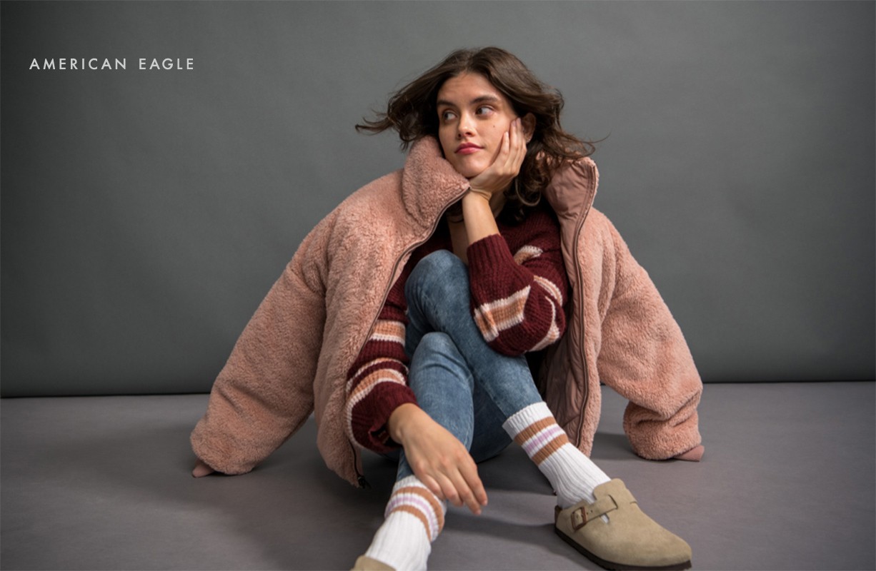 A young woman poses indoors wearing a pink sherpa jacket, striped sweater, jeans, socks, and clogs while sitting on a grey surface.