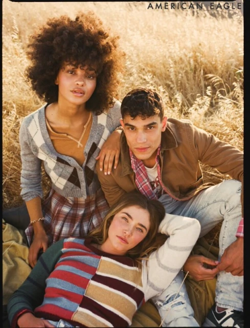Three young adult friends pose together in a field of tall grass, wearing casual sweaters, jeans, and plaids. American Eagle branding visible.