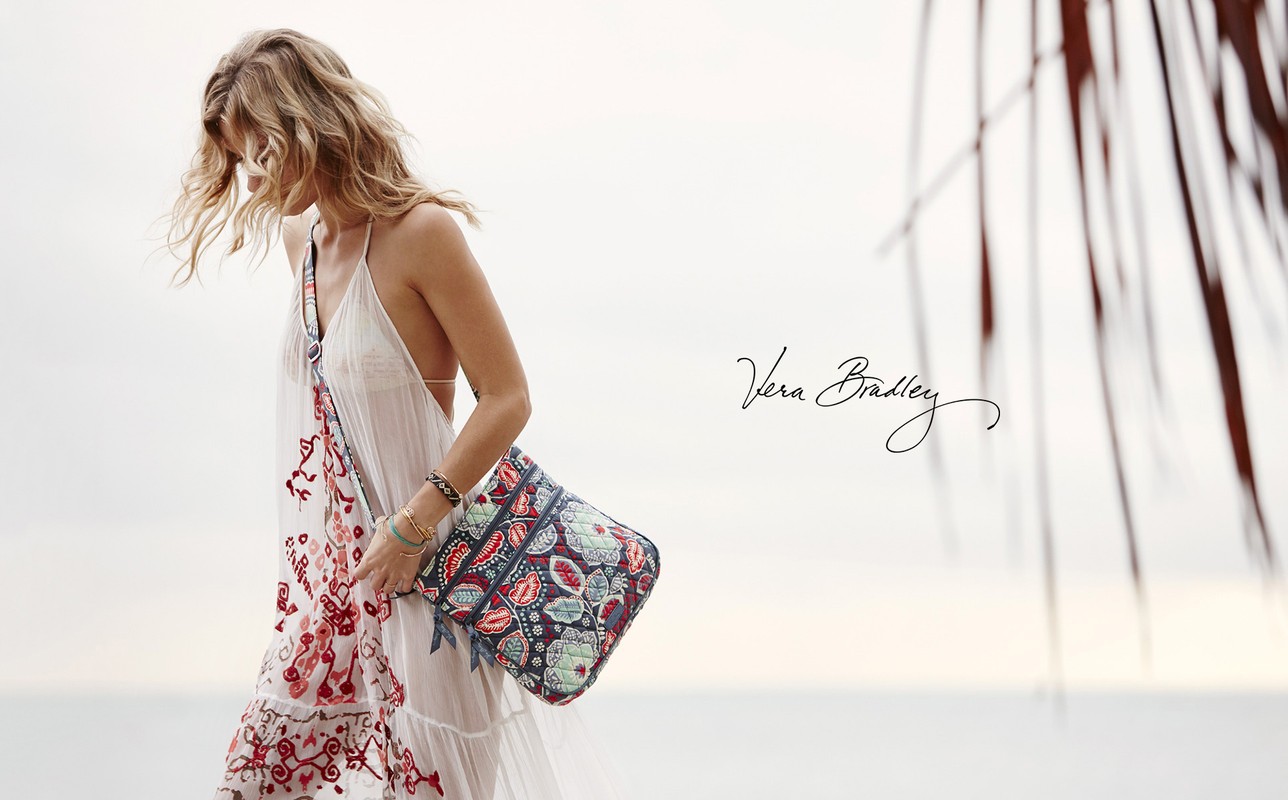 Woman in white summer dress and patterned shoulder bag walking on beach with blurred palm frond. Vera Bradley branding.