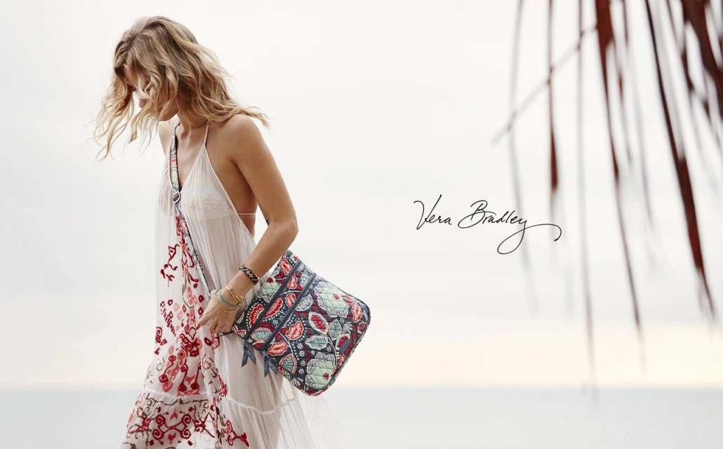 Woman in white summer dress and patterned shoulder bag walking on beach with blurred palm frond. Vera Bradley branding.