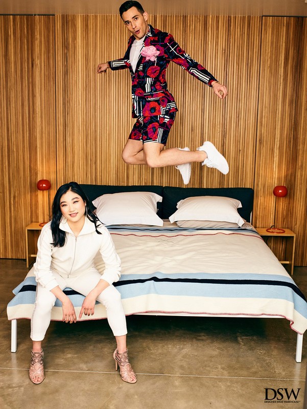 An adult male jumping in the air in front of a bed with an adult female sitting at the edge of the bed in an indoor setting.