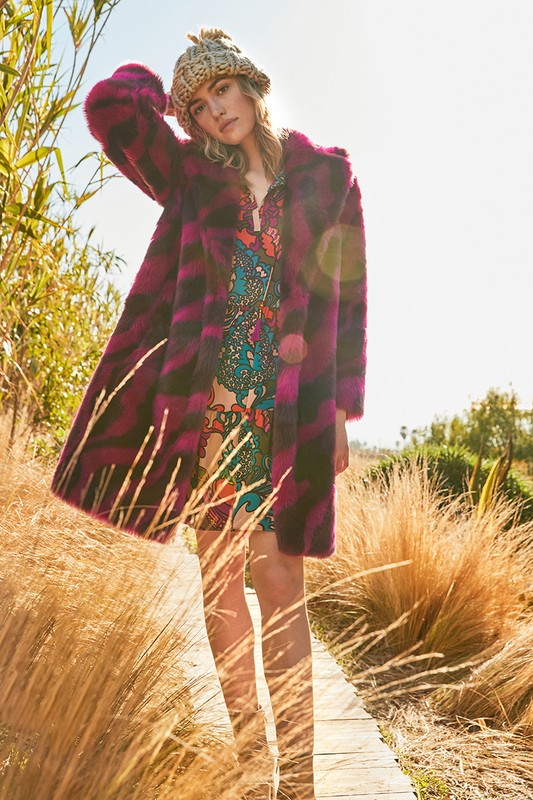A young adult female model poses outdoors wearing a striped faux fur coat, patterned mini dress, and bucket hat on a wooden walkway.