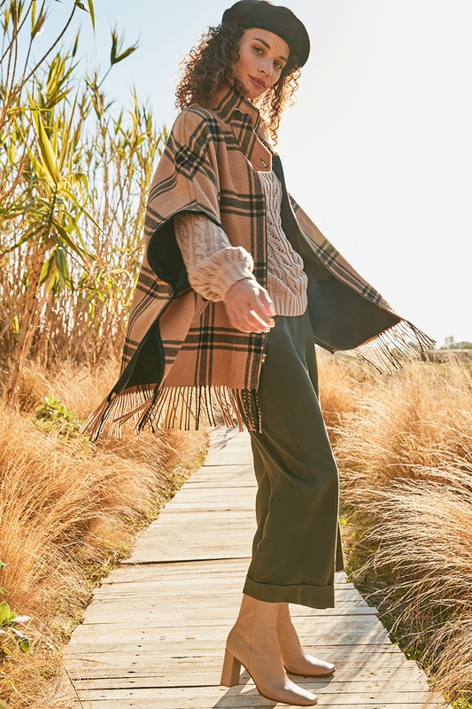 A woman poses on a wooden path in a plaid poncho, sweater, hat, and heeled shoes, surrounded by dry grass and bright, sunny lighting.