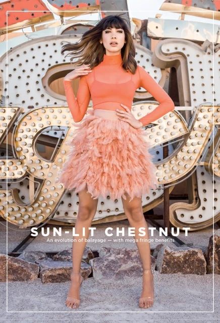 A fashionable woman posing outdoors in a long-sleeved crop top and feathered skirt against a light-bulb sign, showcasing a hair style.