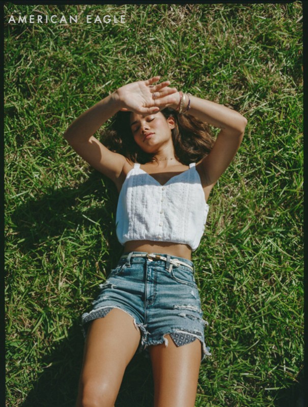 A woman in a white top and denim shorts lies on green grass with her arms raised, blocking the sun. The sky is not visible.
