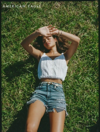 A woman in a white top and denim shorts lies on green grass with her arms raised, blocking the sun. The sky is not visible.