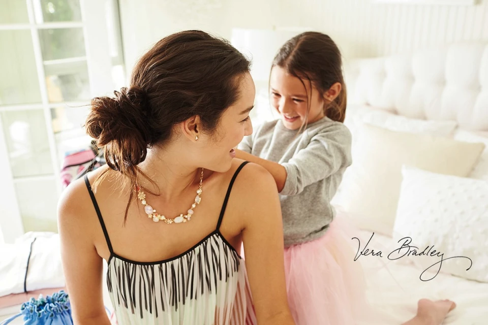 Smiling child in pink tutu fixes necklace on a woman wearing a striped dress in a bright indoor setting with natural light.