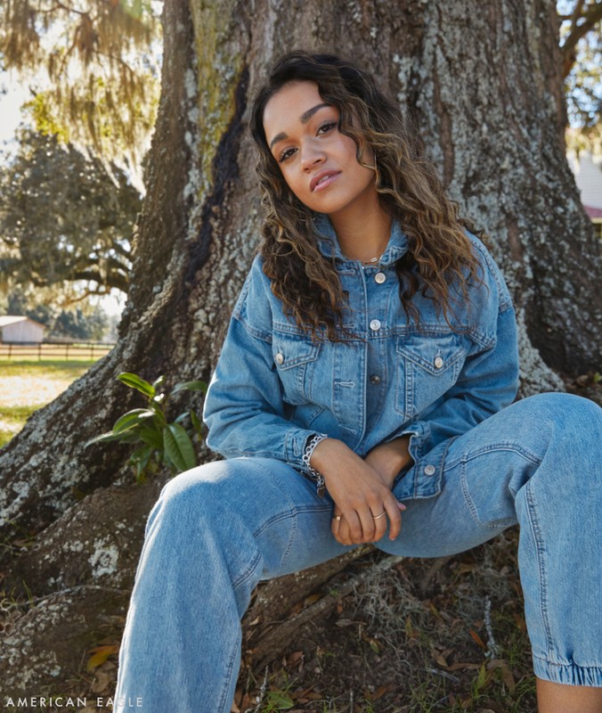 A young woman with curly brown hair sits in front of a large tree wearing a denim jacket and jeans in an outdoor setting.
