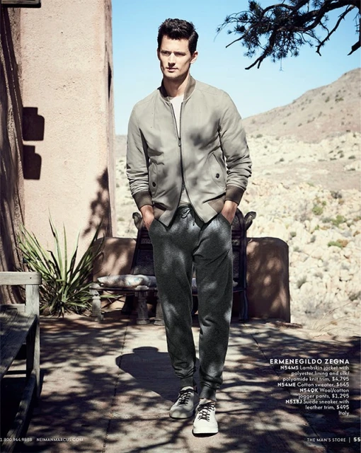Man in grey bomber jacket and sweatpants walks in a sunlit outdoor setting with an adobe building and desert landscape backdrop.