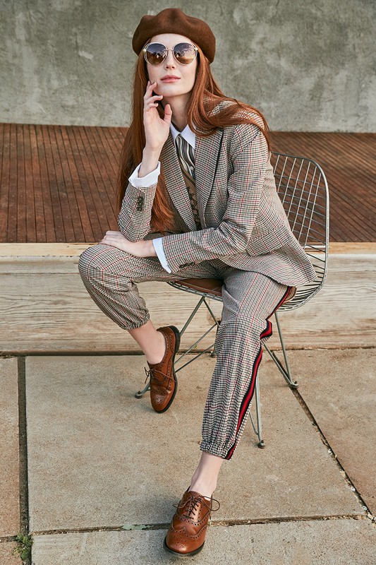 Stylish woman with long red hair sits on a wire chair wearing a brown beret, sunglasses, and patterned suit with Oxford shoes outdoors.