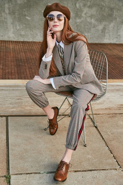 Stylish woman with long red hair sits on a wire chair wearing a brown beret, sunglasses, and patterned suit with Oxford shoes outdoors.