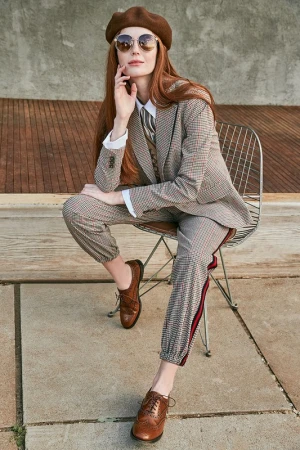 Stylish woman with long red hair sits on a wire chair wearing a brown beret, sunglasses, and patterned suit with Oxford shoes outdoors.