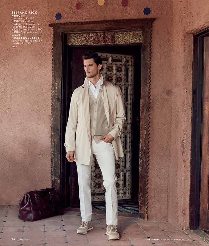 Man in stylish outerwear, vest, white pants poses near an ornately carved doorway with a leather duffel bag at his feet.