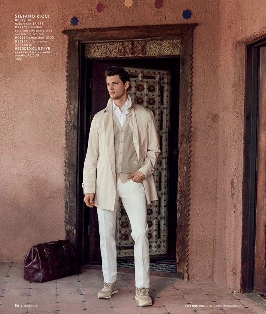 Man in stylish outerwear, vest, white pants poses near an ornately carved doorway with a leather duffel bag at his feet.