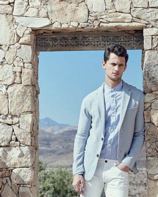 Handsome man in stylish light blue blazer and white pants stands in stone doorway with mountain landscape background under clear sky.