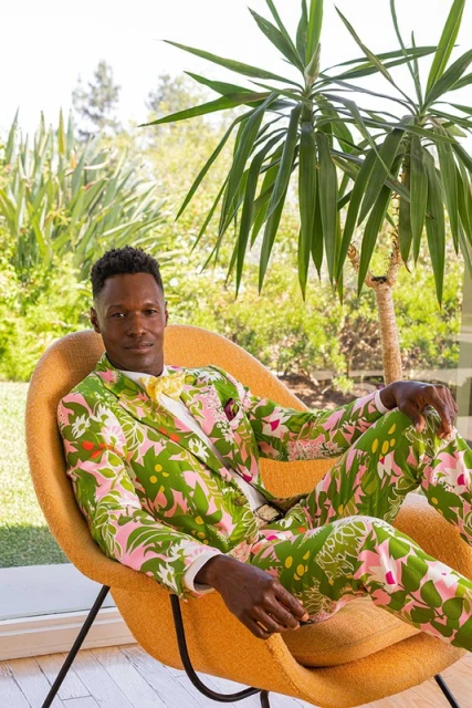 Adult man in patterned green and pink suit reclines in yellow modern chair indoors near large window with outdoor foliage.