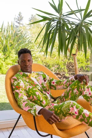 Adult man in patterned green and pink suit reclines in yellow modern chair indoors near large window with outdoor foliage.