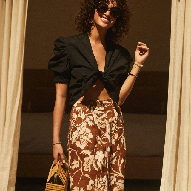 A smiling adult woman in a black tie-front top and floral pants holds a woven purse near a window with a curtain, bathed in soft light.