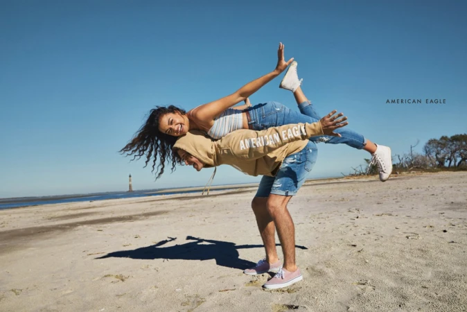 A smiling couple performs a partner yoga pose on a beach under a clear blue sky. The woman lies atop the man, both wearing casual clothing.
