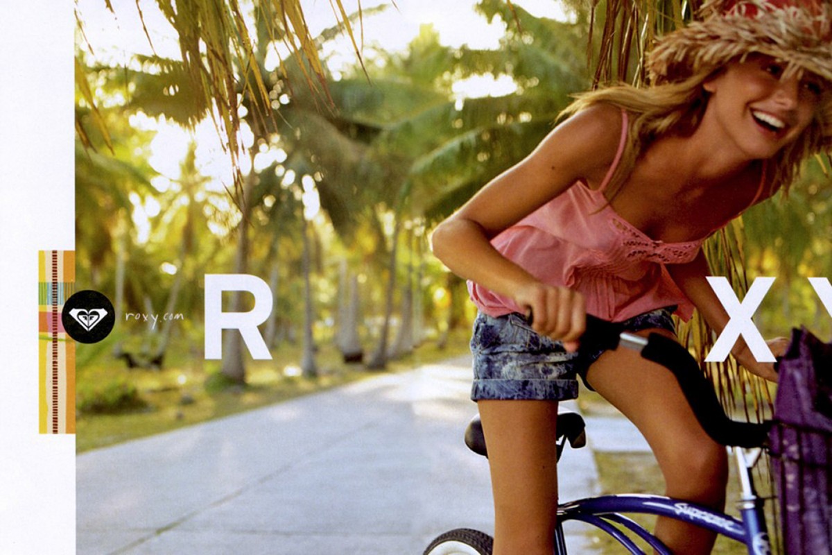 Smiling woman in straw hat rides bicycle on tropical road with palm trees in sunny outdoor shot for a fashion advertisement.