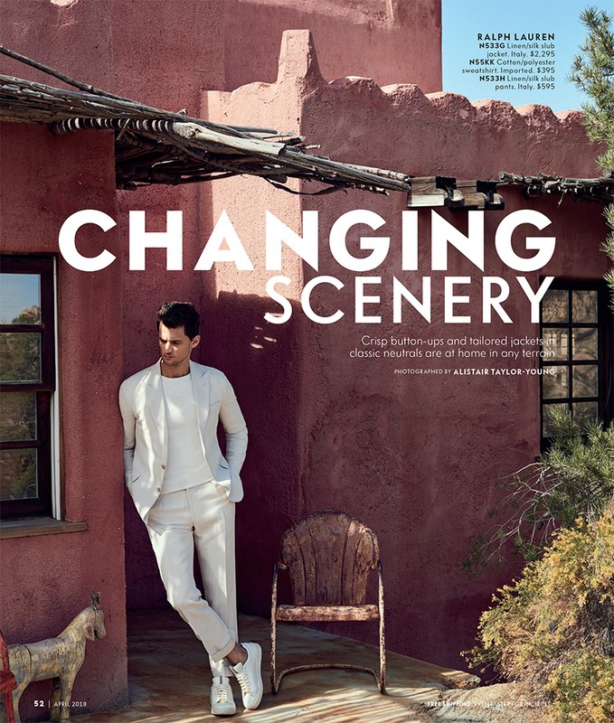 Male fashion model in an off-white suit stands in front of a terracotta-colored building on a sunny day with an antique wooden chair.
