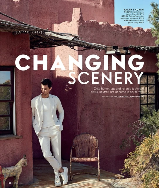 Male fashion model in an off-white suit stands in front of a terracotta-colored building on a sunny day with an antique wooden chair.
