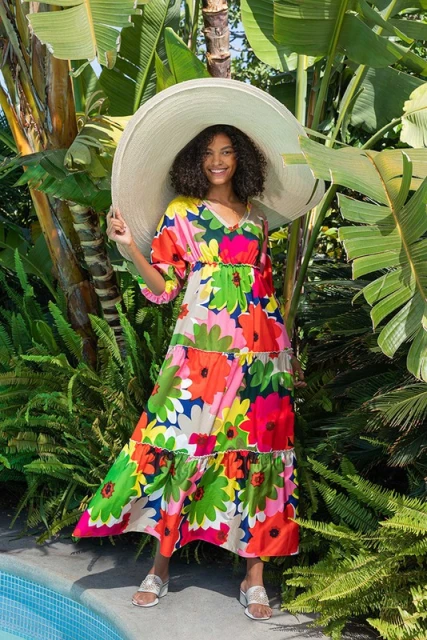 Woman in a floral dress and oversized hat stands near a pool surrounded by tropical foliage, smiling brightly into the camera.