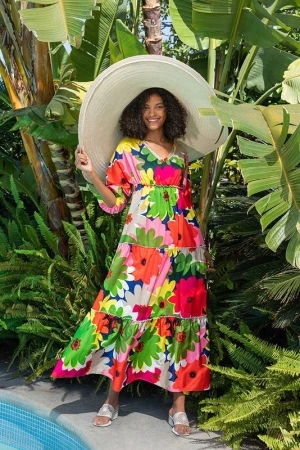 Woman in a floral dress and oversized hat stands near a pool surrounded by tropical foliage, smiling brightly into the camera.